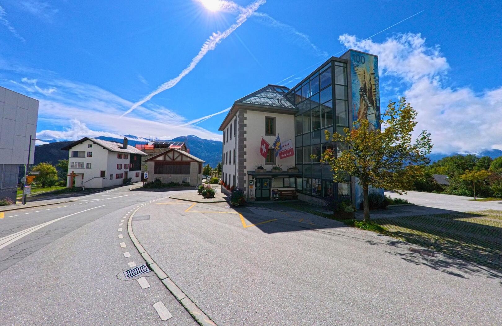 Das Gemeindehaus von Ayent (1966, Kanton Wallis) mit Glasfassade und aufgehängten Fahnen steht an einer Strassenkurve vor alpiner Bergkulisse.