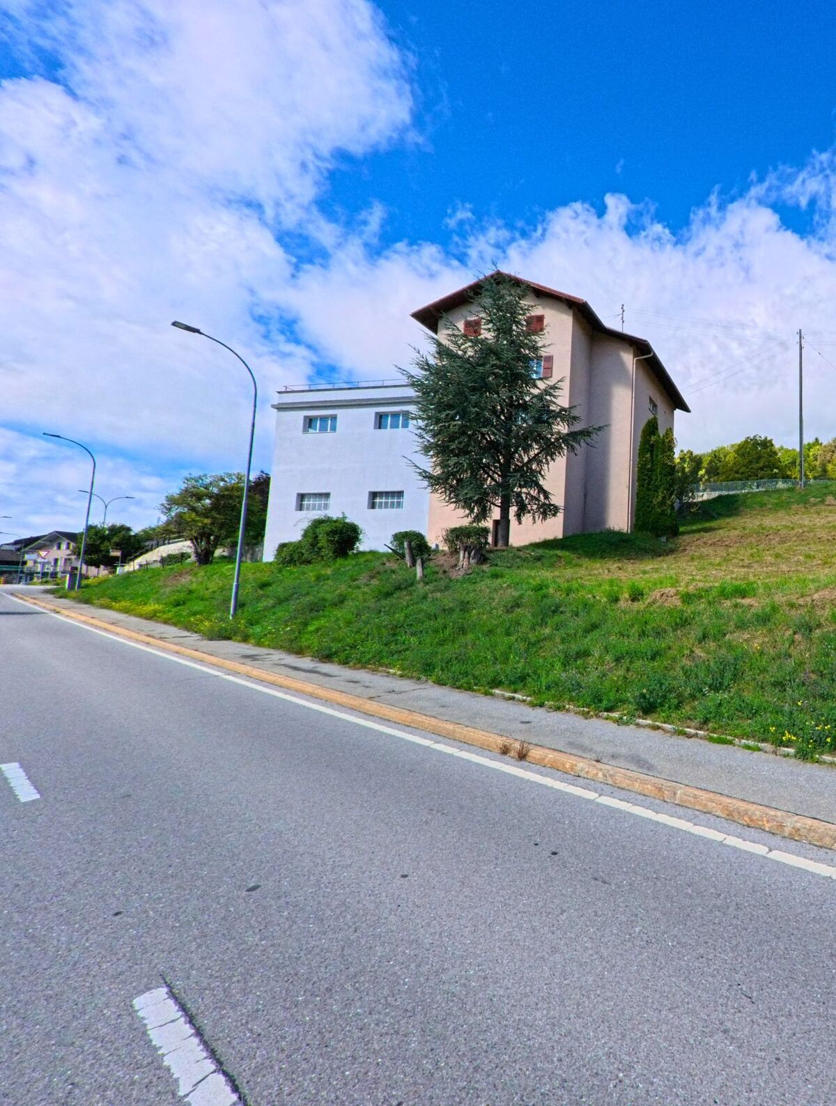 Ein Wohnhaus mit hellem Anstrich steht oberhalb einer Strasse in Ayent (1966, Kanton Wallis) neben einer Wiese und einzelnen Baeumen.