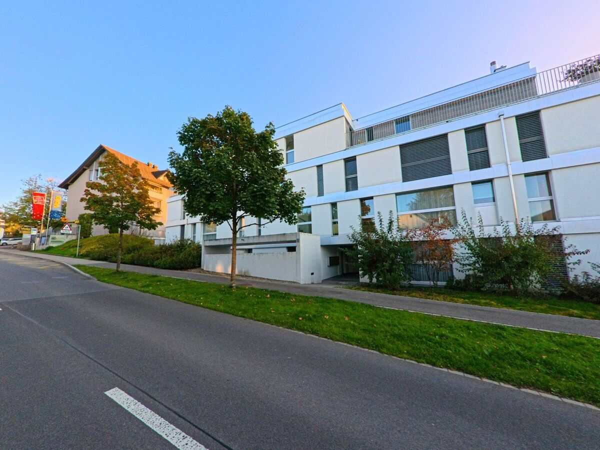 In Baar (6319, Kanton Zug) stehen moderne Wohngebaeude mit klaren Linien entlang einer leicht ansteigenden Strasse, umgeben von Baeumen und Strauchwerk.