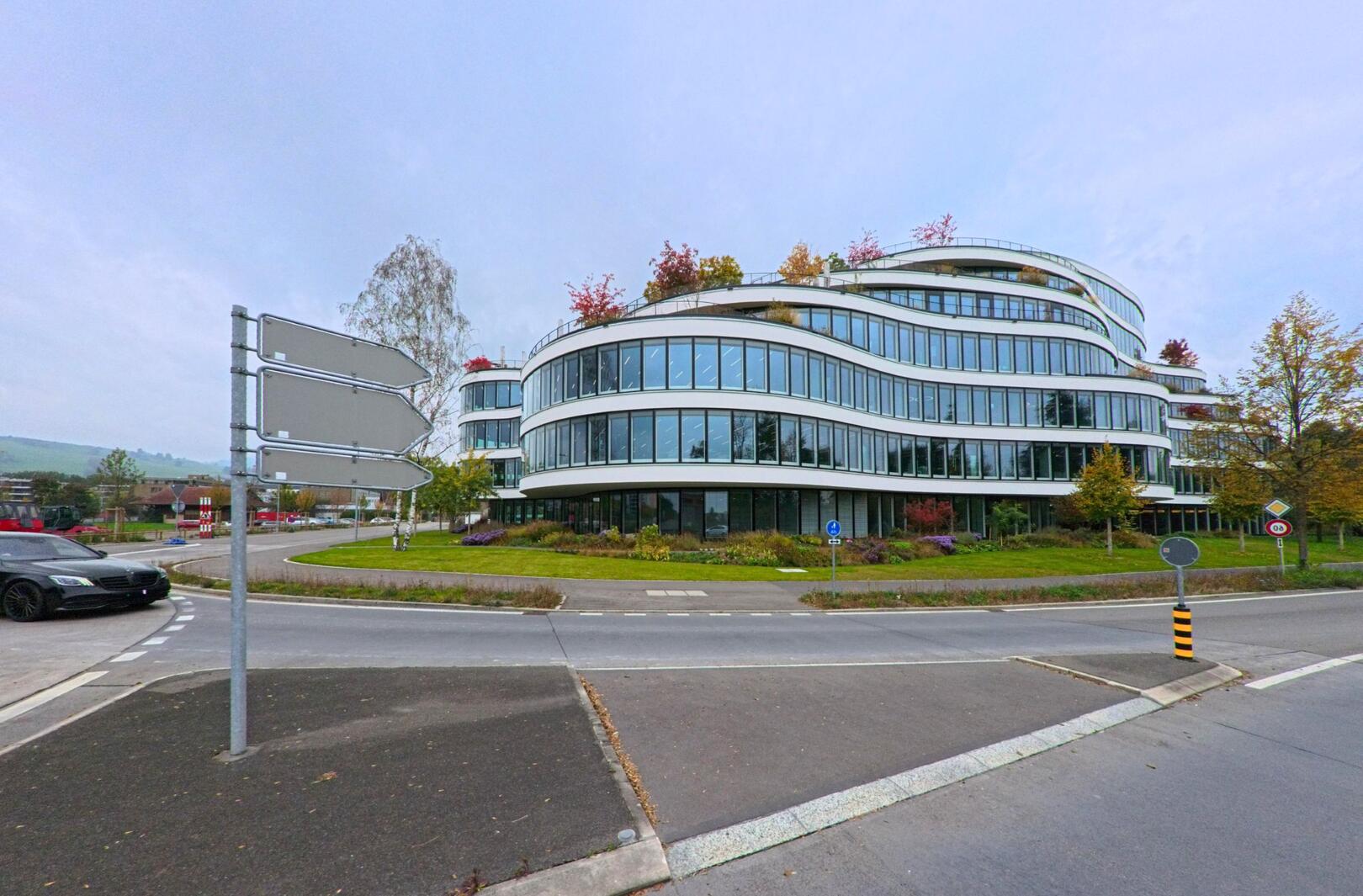 In Baar (6319, Kanton Zug) steht ein modernes, geschwungenes Buerohaus mit grossen Glasfronten an einem Kreisverkehr, umgeben von herbstlicher Bepflanzung.
