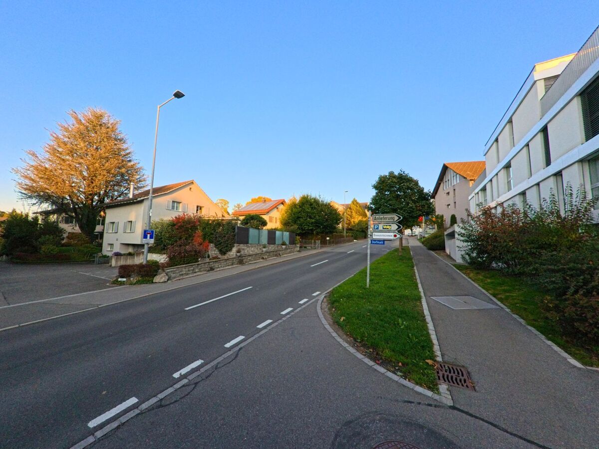 Eine leicht ansteigende Strasse in Baar (6319, Kanton Zug) fuehrt zwischen Wohnhaeusern und einem modernen Gebaeude hindurch, begleitet von Wegweisern.