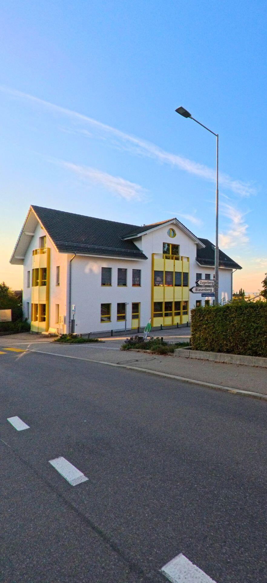 In Baar (6319, Kanton Zug) steht ein weiss-gelbes Gebaeude mit markanten Fensterfronten an einer ruhigen Strasse im warmen Abendlicht.