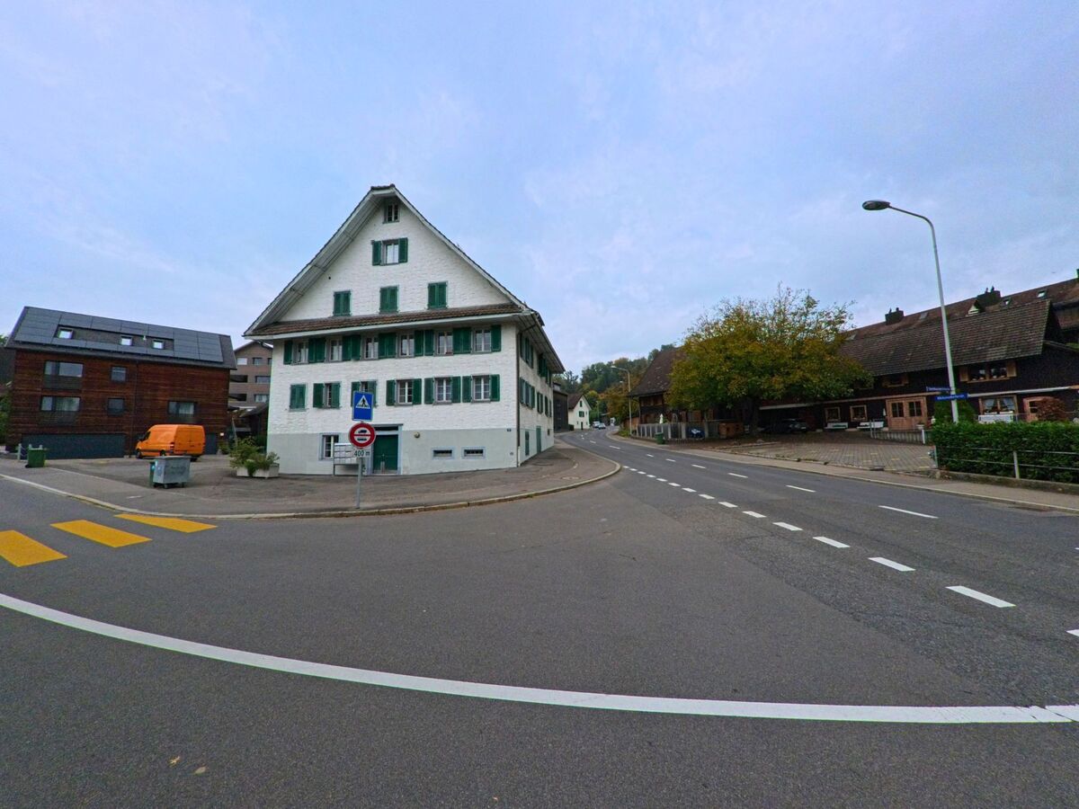 In Baar (6319, Kanton Zug) steht ein weisses Haus mit gruenen Fensterlaeden an einer breiten Kreuzung, umgeben von traditionellen Gebäuden.