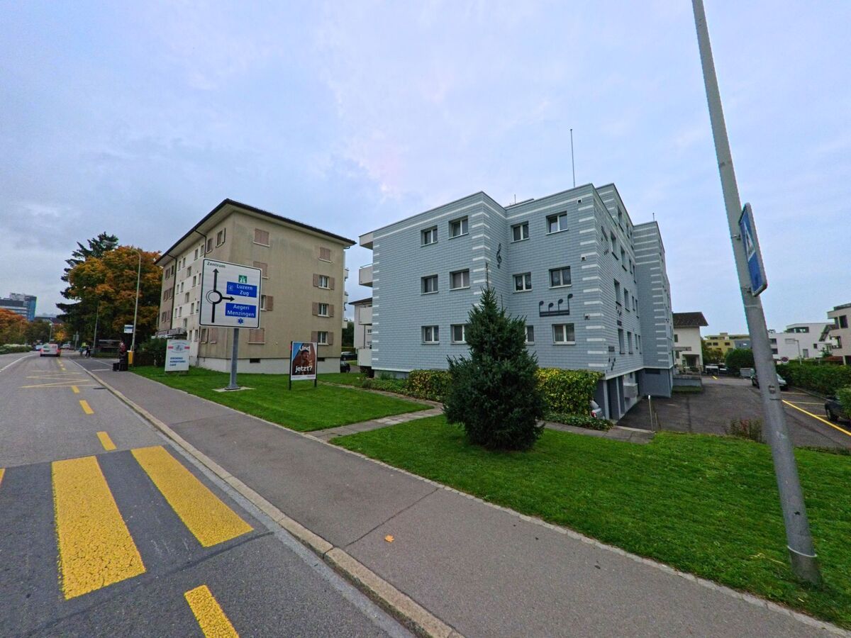 In Baar (6319, Kanton Zug) stehen mehrere Wohnbloecke entlang einer breiten Strasse mit gelben Markierungen und Wegweisern.