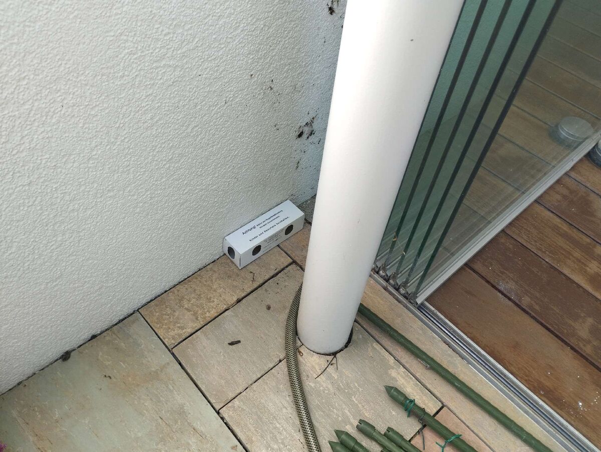 Auf einem Balkon in Bachenbülach (8184, Kanton Zürich) liegt eine Köderstation an der Wand zwischen einer weissen Säule und einer Glasschiebetür.