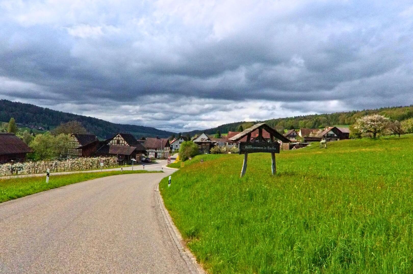 Ein Schild am Ortseingang begrüsst Besucher in Bachs im Kanton Zürich, PLZ 8164, mit Blick auf die Fachwerkhäuser des Dorfkerns unter einem bewölkten Himmel.
