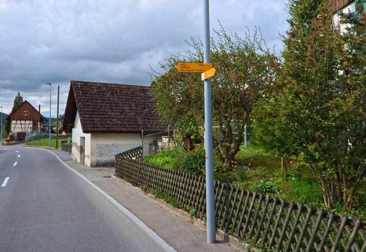 Ein gelber Wanderwegweiser an der Hauptstrasse von Bachs im Kanton Zürich, PLZ 8164, zeigt Richtungen nach Schöfflisdorf und Oberweningen inmitten einer grünen Gartenumgebung.