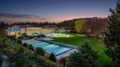 In Schinznach-Bad (5116, Kanton Aargau) leuchten die Aussenbecken der Bad Schinznach AG am Abend in warmem Licht, umgeben von gepflegtem Gruen und herbstlichen Baeumen.