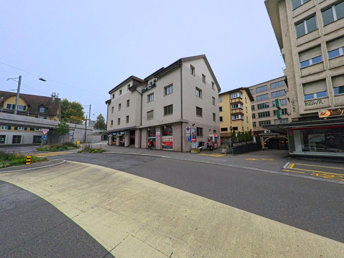 Ein Eckgebaeude mit Geschaeften im Erdgeschoss steht in Baden (5400, Kanton Aargau) an einer Strasse mit Fussgaengerueberweg und angrenzenden modernen Bauten.