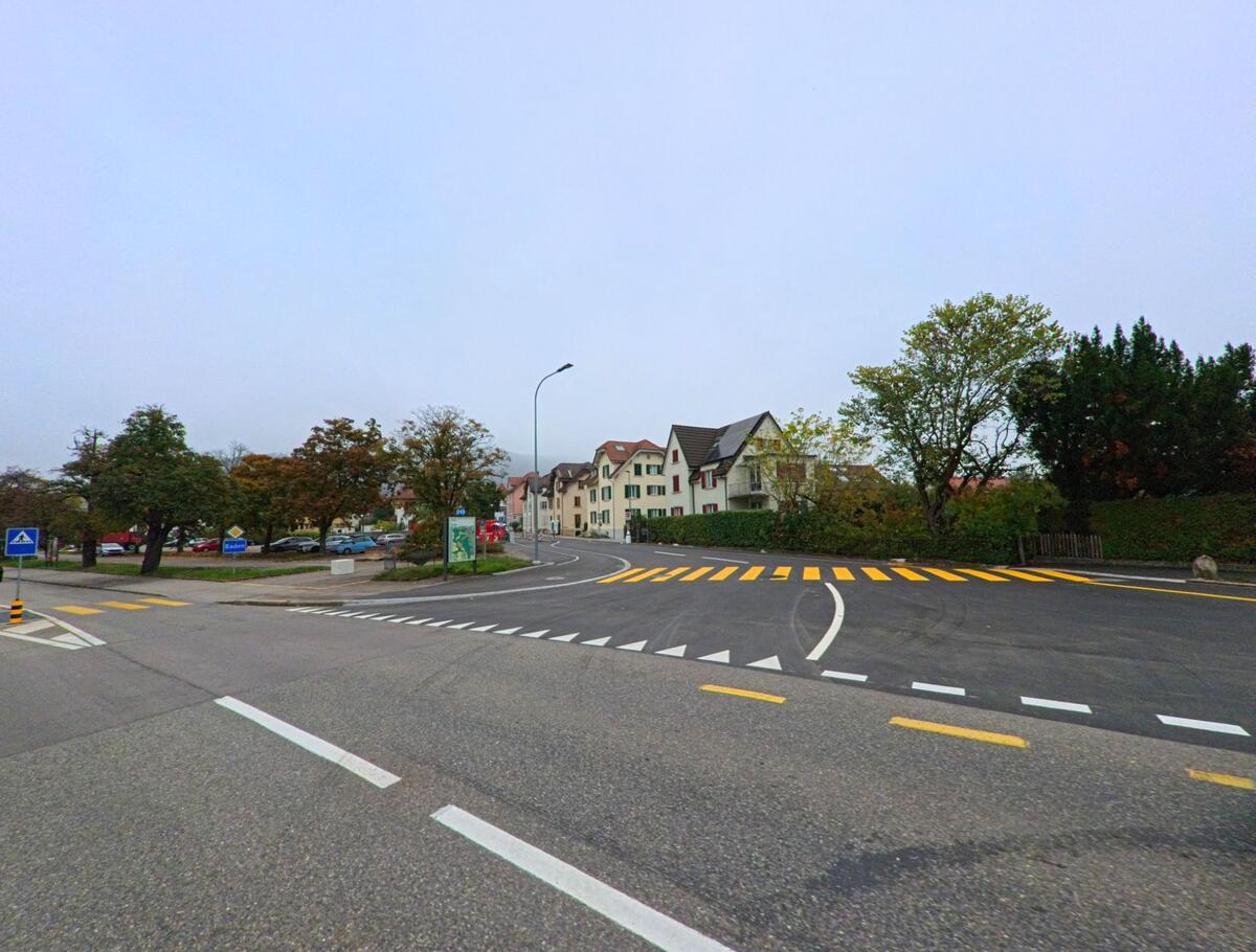 Eine breite Kreuzung in Baden (5400, Kanton Aargau) fuehrt vorbei an Wohnhaeusern und Baeumen, mit klar markierten Fussgaengerstreifen und einer leicht gekruemmten Strasse.