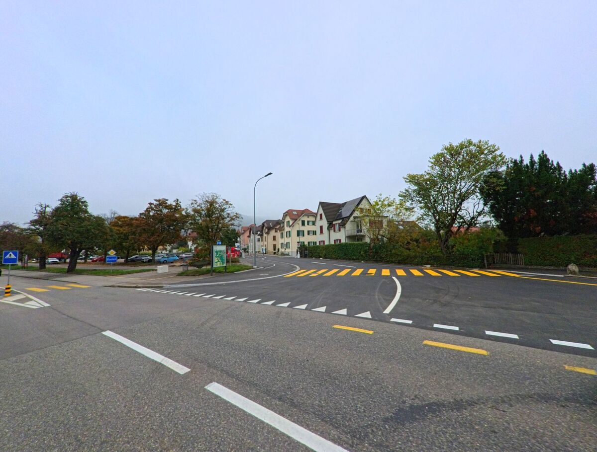 Eine breite Kreuzung in Baden (5400, Kanton Aargau) fuehrt vorbei an Wohnhaeusern und Baeumen, mit klar markierten Fussgaengerstreifen und einer leicht gekruemmten Strasse.