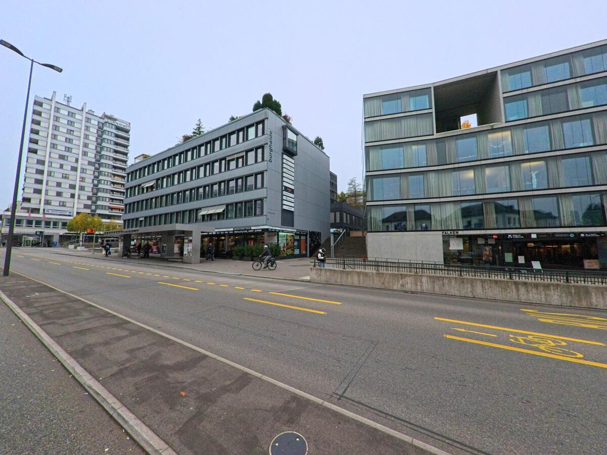 Moderne Wohn- und Geschaeftsgebaeude mit grossen Glasfassaden saeumen in Baden (5400, Kanton Aargau) eine belebte Strasse mit Radfahrer und Passanten.