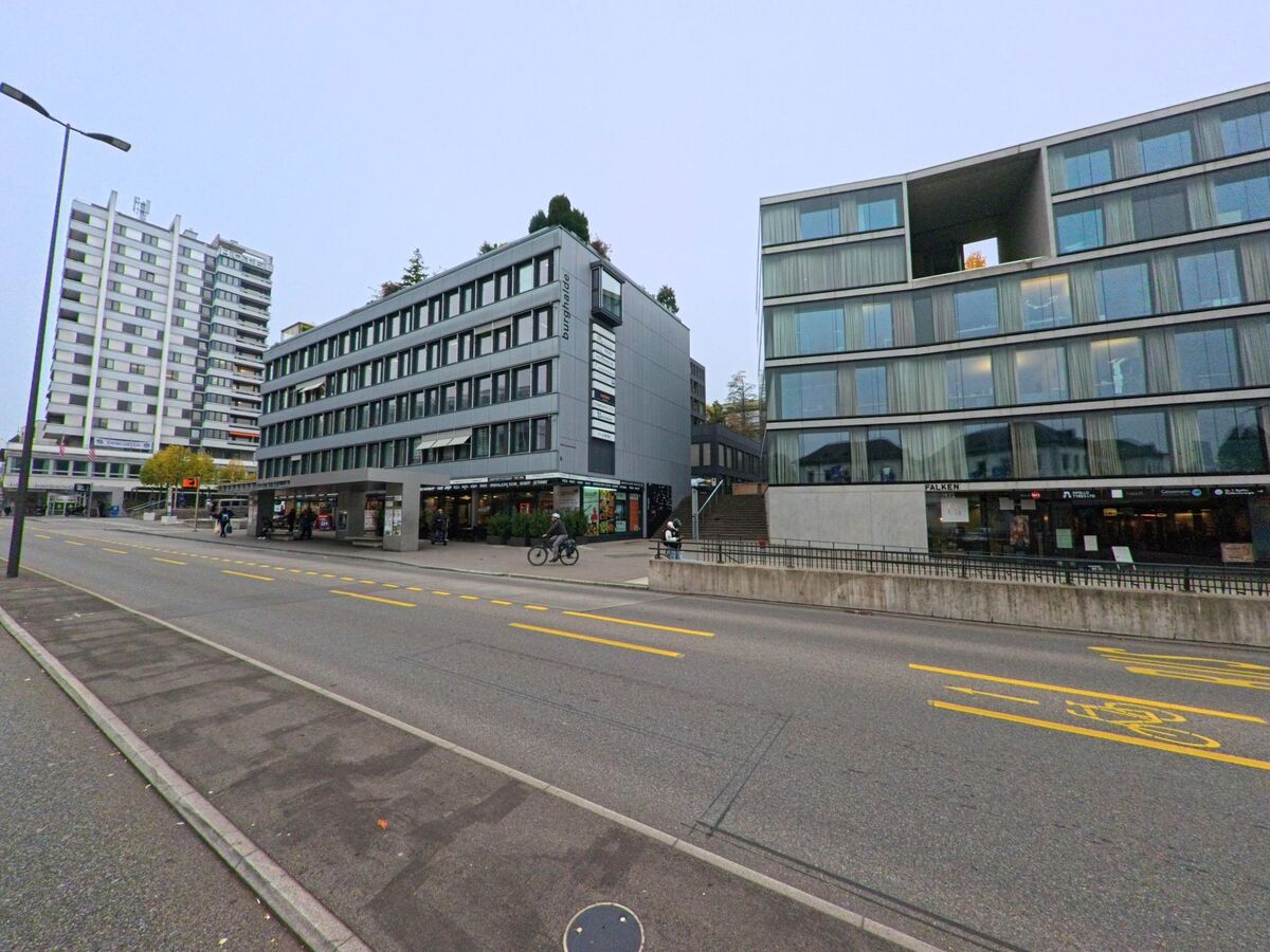 Moderne Wohn- und Geschaeftsgebaeude mit grossen Glasfassaden saeumen in Baden (5400, Kanton Aargau) eine belebte Strasse mit Radfahrer und Passanten.