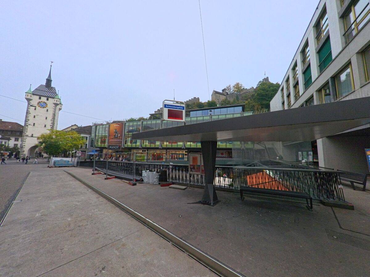 Am Schlossbergplatz in Baden (5400, Kanton Aargau) stehen moderne Gebaeude neben dem markanten Stadtturm, waehrend im Hintergrund die Schlossruine auf dem Huegel zu sehen ist.