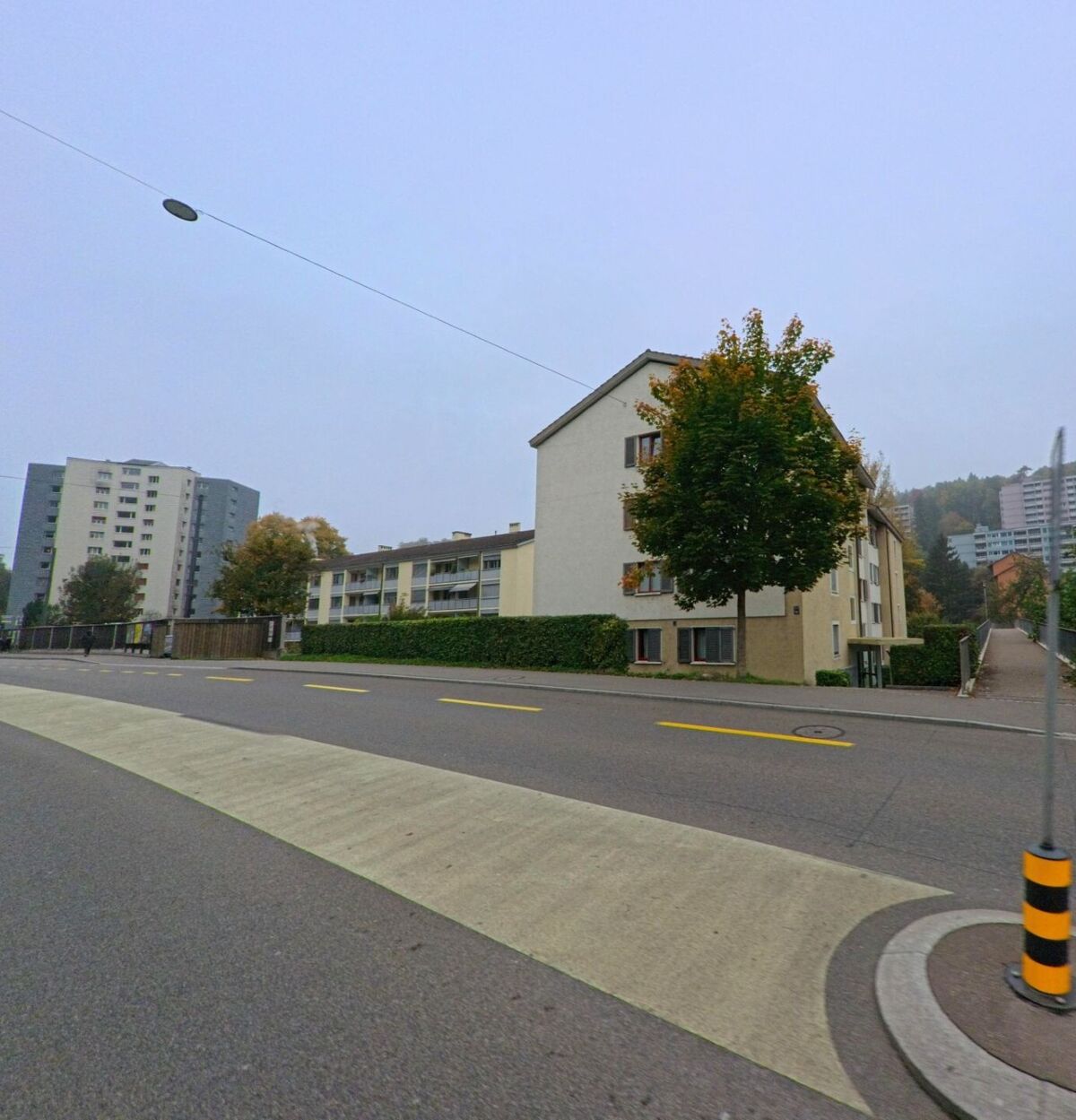 Mehrgeschossige Wohnhaeuser und ein einzeln stehender Baum saeumen in Baden (5400, Kanton Aargau) eine ruhige Strasse mit breitem Fussgaengerstreifen und Asphaltbelag.