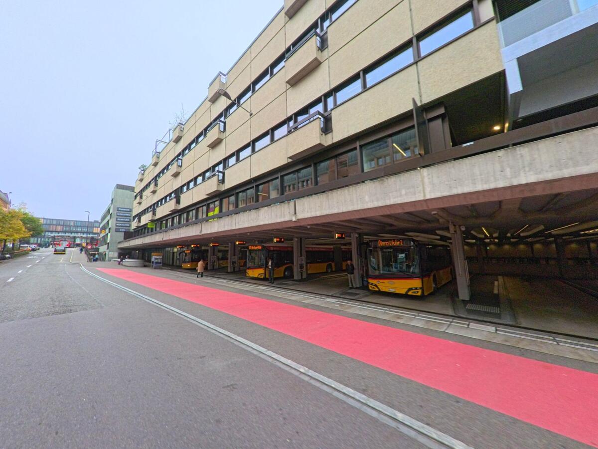 Mehrere gelbe Postautos stehen an der Bushaltestelle unter einem grossen Betongebaeude in Baden (5401, Kanton Aargau) entlang einer Strasse mit rotem Busstreifen.
