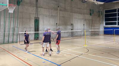 Vier Personen stehen mit Schlaegern am Badmintonnetz in einer Turnhalle in Muttenz (4132, Kanton Baselland) nach einem gespielten Ballwechsel zusammen.