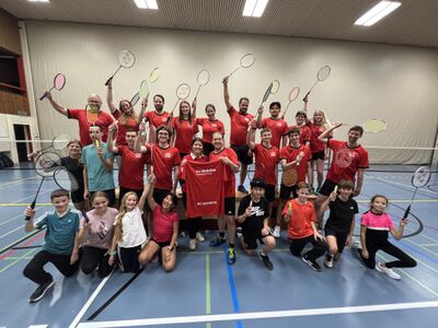 Gruppenfoto des Badmintonclubs Schwyz 6430 im Kanton Schwyz mit Kindern, Jugendlichen und Erwachsenen in der Turnhalle, rote Trikots, erhobene Schläger und gemeinsamer Teamgeist.
