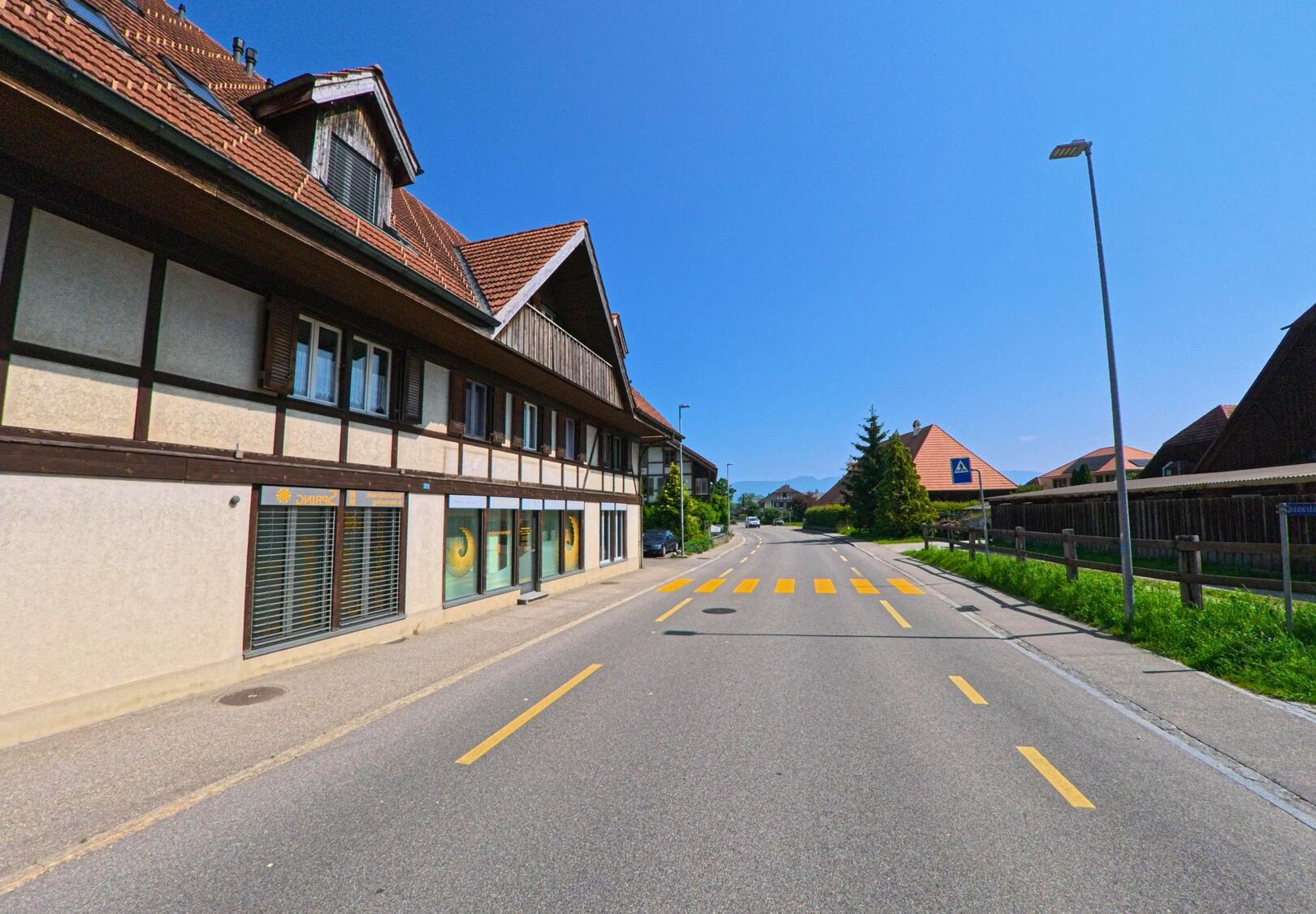 In Bätterkinden (3315, Kanton Bern) verläuft eine ruhige Quartierstrasse mit Fussgängerstreifen entlang traditioneller Wohnhäuser unter strahlend blauem Himmel.
