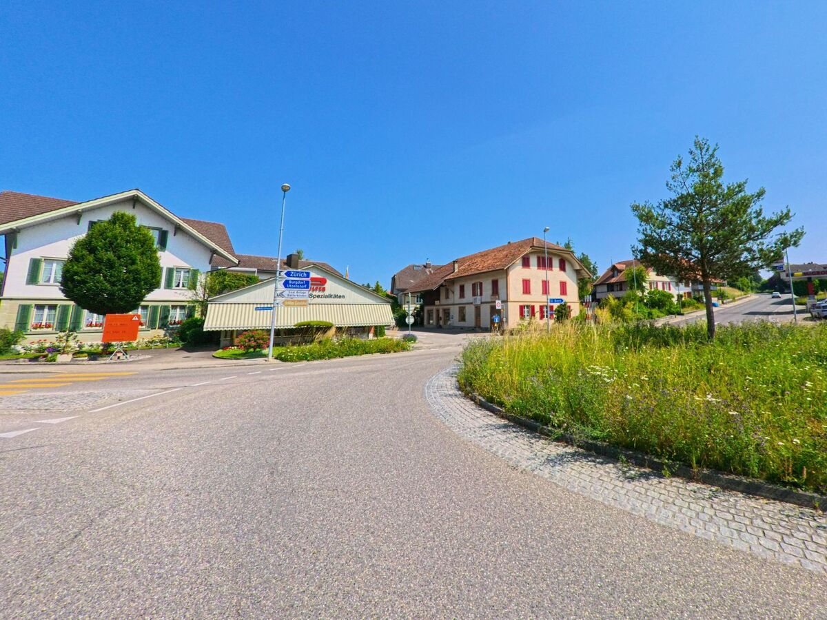 In Bätterkinden (3315, Kanton Bern) verzweigt sich die Hauptstrasse an einer kleinen Verkehrsinsel mit Wegweisern und angrenzenden Wohn- und Geschäftshäusern.