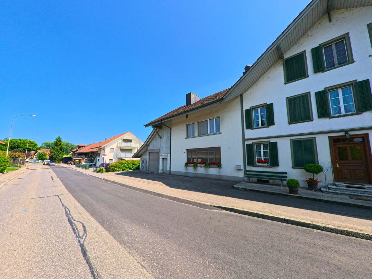 In Bätterkinden (3315, Kanton Bern) säumen gepflegte Wohnhäuser mit Fensterläden und Blumenkästen eine ruhige Strasse unter wolkenlosem Himmel.