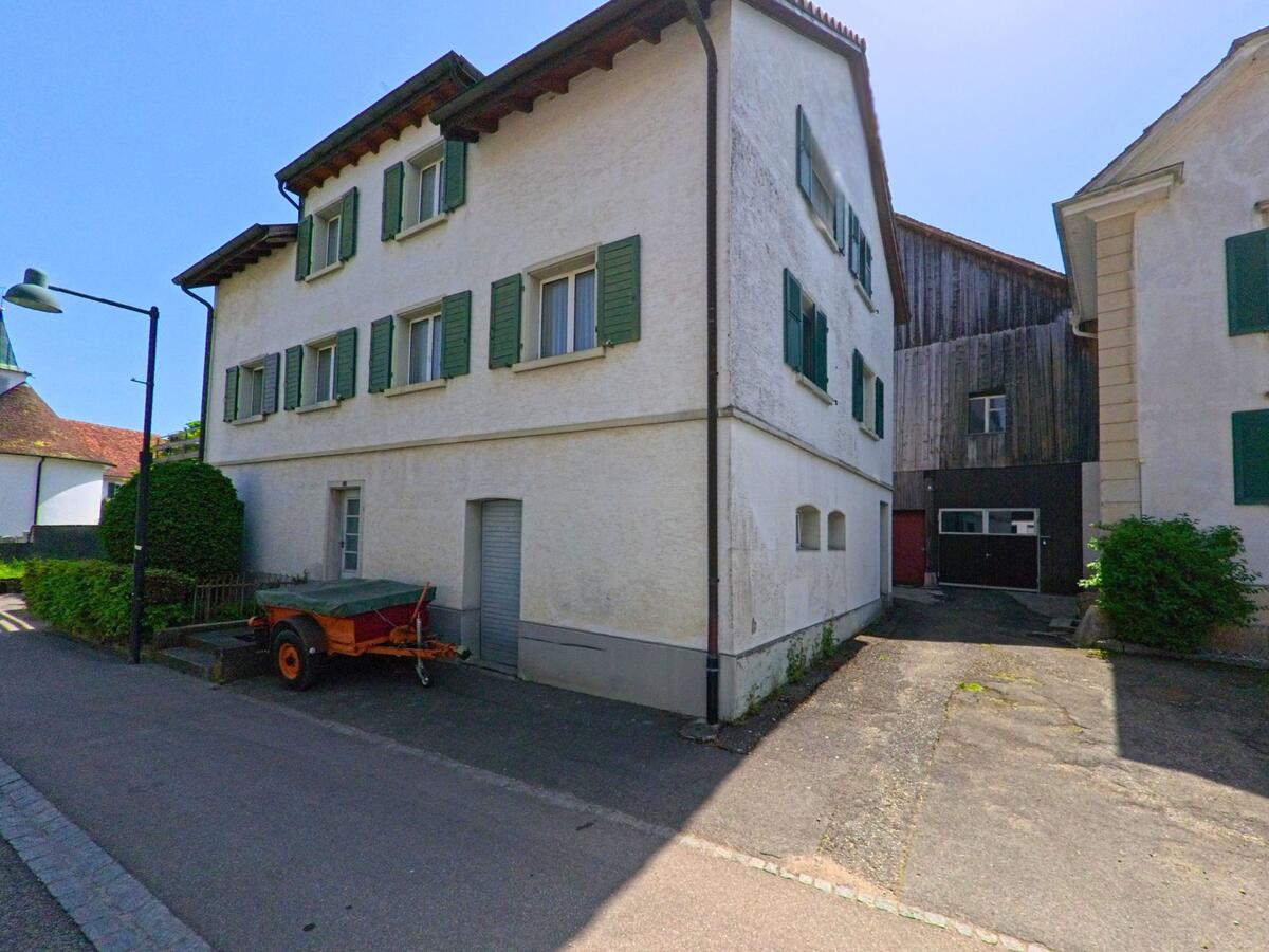 In Bättwil im Kanton Solothurn, PLZ 4112, stehen zwei ältere Wohnhäuser mit grünen Fensterläden eng beieinander, dazwischen ein schmaler Durchgang zu einem Holzbau.