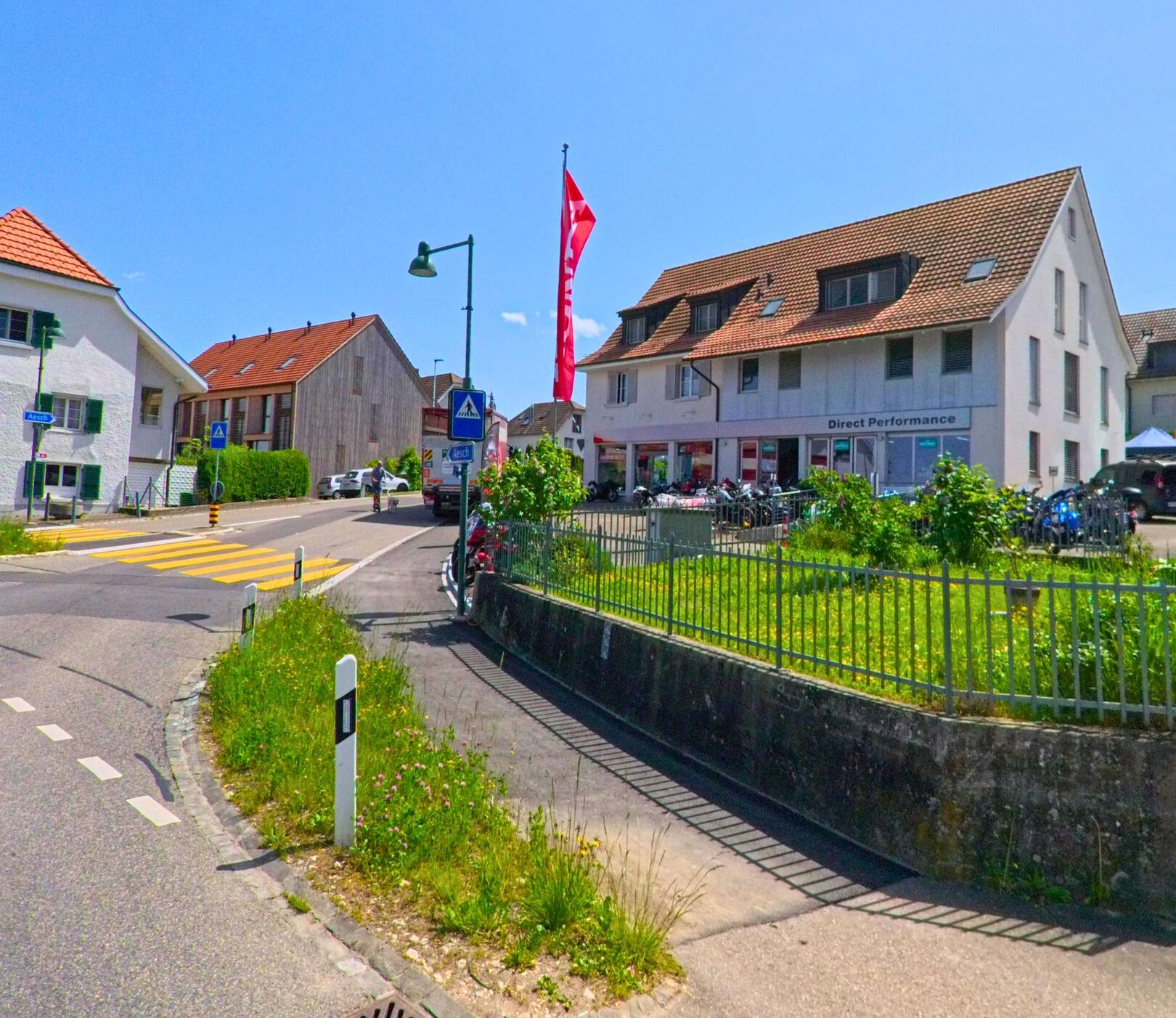 In Bättwil im Kanton Solothurn, PLZ 4112, liegt eine zentrale Kreuzung mit Zebrastreifen, Geschäftshaus und angrenzenden Wohnhäusern.