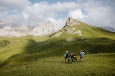 Drei Mountainbiker halten in Adelboden (3715, Kanton Bern) auf einer gruene Wiese mit Blick auf felsige Gipfel und sanfte Alpenhaenge an.