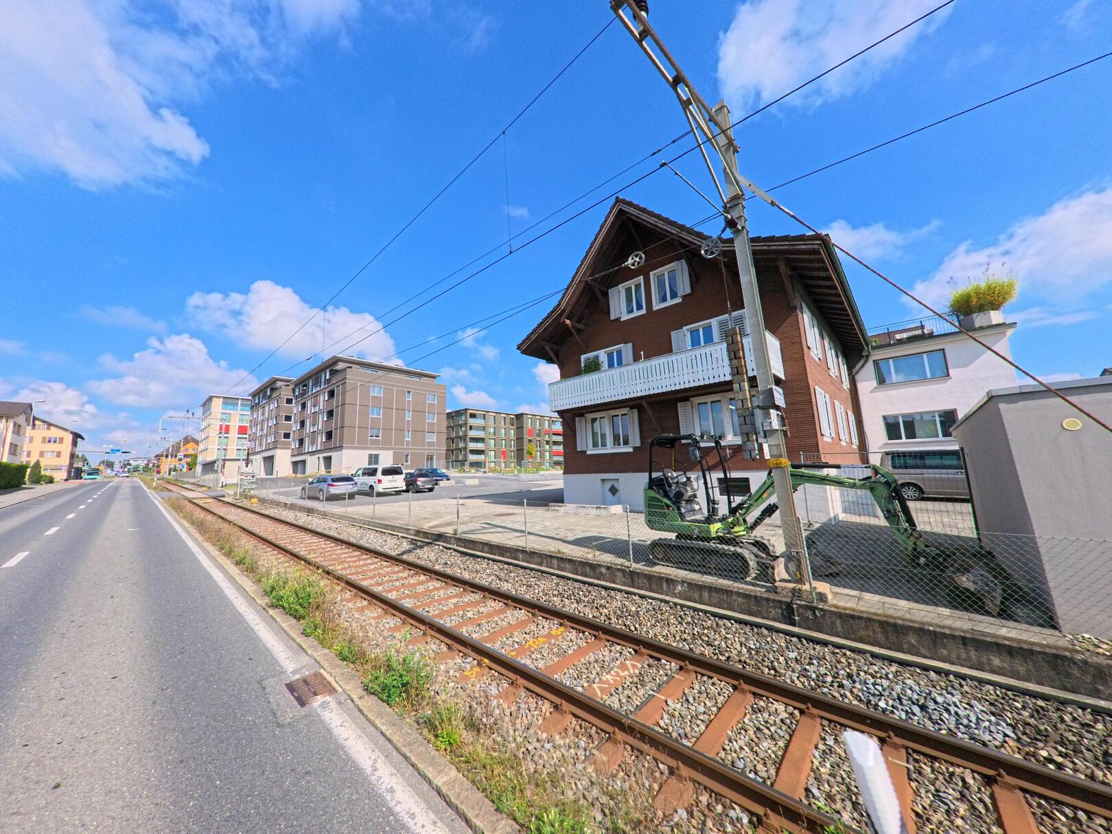 Entlang der Bahnlinie in Ballwil (6275, Kanton Luzern) stehen moderne Wohnhaeuser neben einem traditionellen Holzhaus mit Balkon.