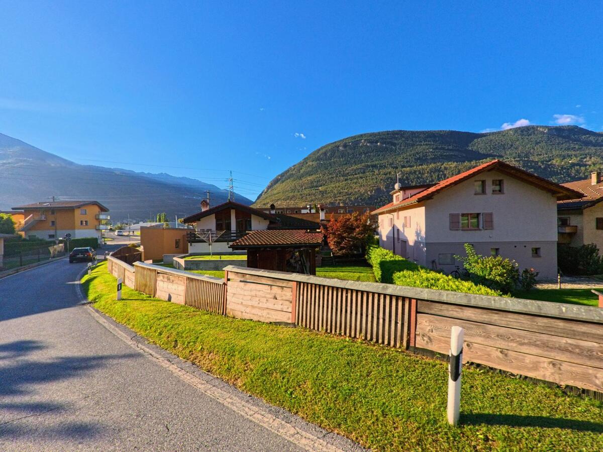 Einfamilienhäuser mit Gärten und Holzzäunen liegen in Baltschieder (3937, Kanton Wallis) am Rand einer Strasse mit Blick auf die bewaldeten Berge.