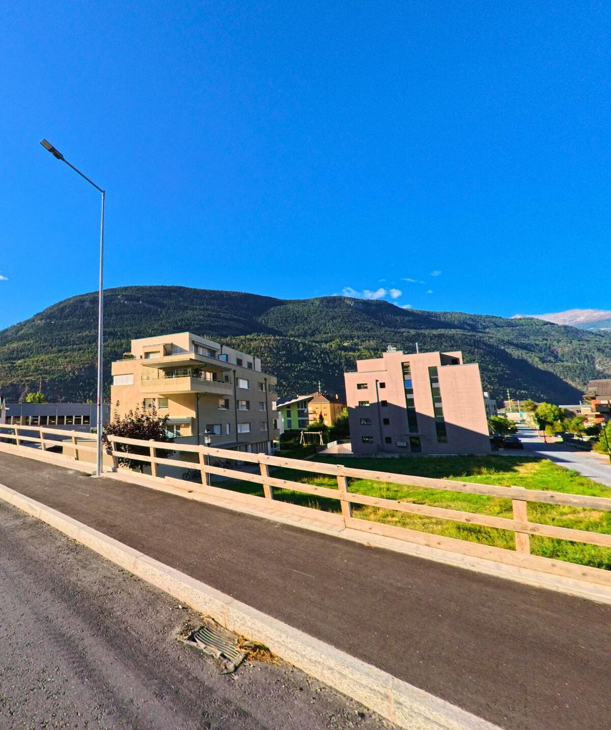 In Baltschieder (3937, Kanton Wallis) stehen moderne Wohngebäude am Strassenrand, dahinter erhebt sich ein bewaldeter Berg.