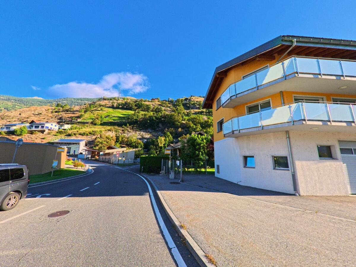 Ein modernes Haus mit hellgelber Fassade und Balkonen steht in Baltschieder (3937, Kanton Wallis) an einer geschwungenen Strasse unterhalb der Hügel.