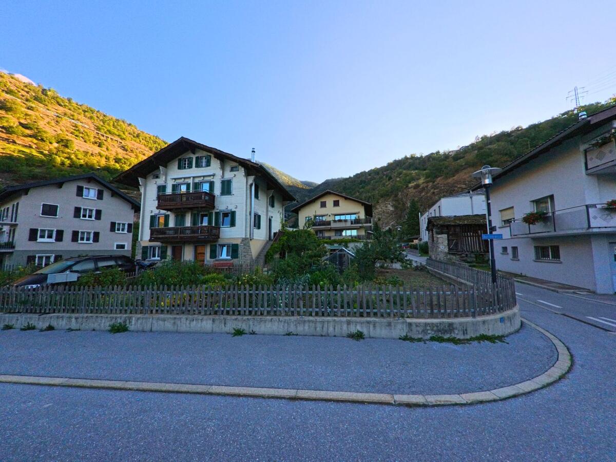 Mehrere Wohnhäuser mit Gärten und Holzzäunen liegen in Baltschieder (3937, Kanton Wallis) am Fuss eines bewaldeten Hügels.