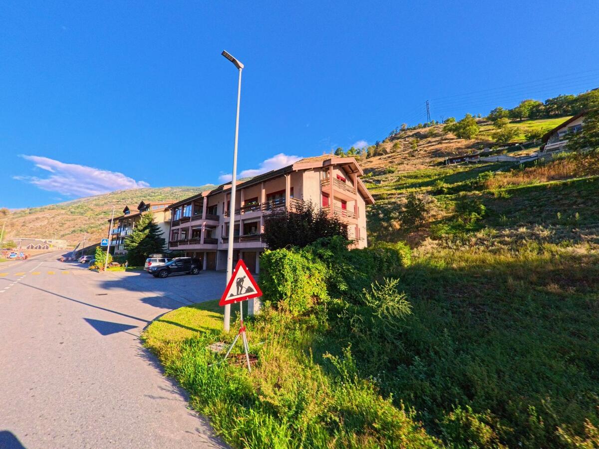 Ein Wohnhaus mit Balkonen liegt in Baltschieder (3937, Kanton Wallis) direkt am Hang oberhalb der Strasse, daneben führt die Fahrbahn ins Dorf.