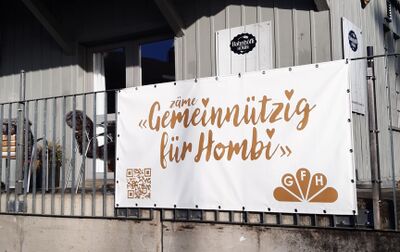 In Hombrechtikon (8634, Kanton Zuerich) haengt ein grosses Banner mit der Aufschrift „zaeme Gemeinnuetzig fuer Hombi“ am Gelaender vor einem Gebaeude.