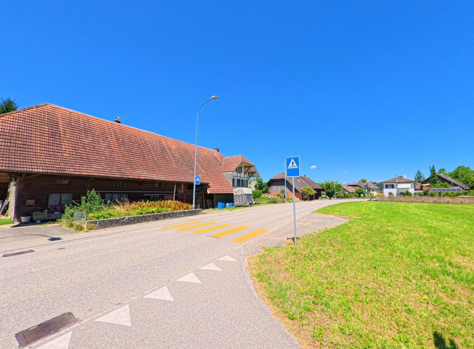 Ein Fussgaengerstreifen fuehrt in Bannwil (4913, Kanton Bern) an einem traditionellen Bauernhaus mit Ziegeldach und einer grossen Wiese vorbei in ein Wohnquartier.