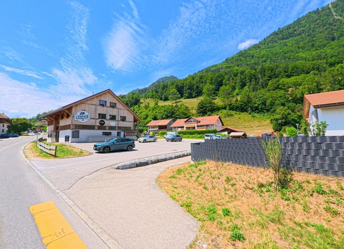 In Bas-Intyamon (1666, Kanton Freiburg) liegt ein Gewerbegebäude mit Parkplatz am Fuss eines bewaldeten Hanges, umgeben von weiteren Häusern und einer kurvigen Strasse.