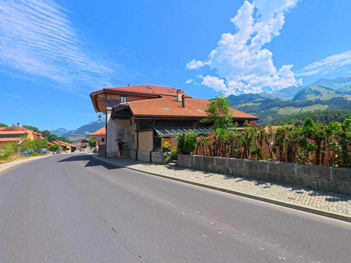 In Bas-Intyamon (1666, Kanton Freiburg) verläuft eine Strasse an einem grossen Wohnhaus mit roten Ziegeldächern entlang, dahinter öffnet sich der Blick auf eine weite Berglandschaft.