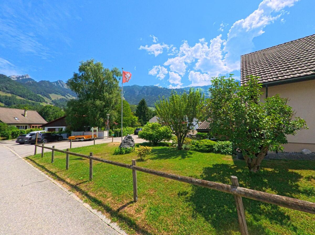 In Bas-Intyamon (1667, Kanton Freiburg) steht eine Fahne auf einer gepflegten Grünfläche zwischen Einfamilienhäusern, mit Blick auf die umliegenden Berge.