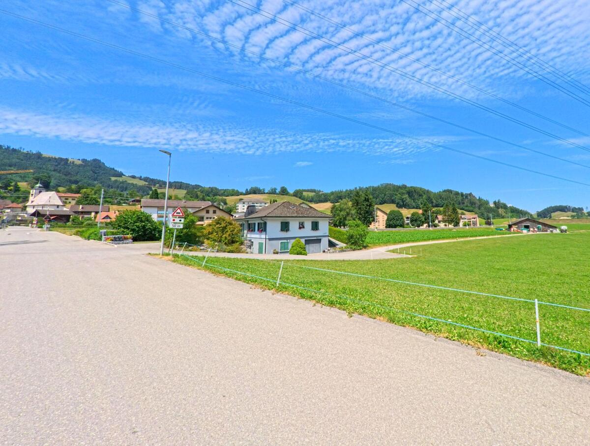 In Bas-Intyamon (1667, Kanton Freiburg) liegt ein einzelnes Wohnhaus zwischen offener Wiese und dem nahen Dorfzentrum mit Blick auf sanfte Hügel und Stromleitungen am Himmel.