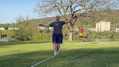 Barfuss gehender Slackliner balanciert über gespannte Slackline auf einer Wiese am Rhein, weitere Person übt im Hintergrund bei frühlischer Stimmung in Basel (4000, Kanton Basel-Stadt).