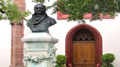 In Basel (4052, Kanton Basel-Stadt) steht die Bueste von J. P. Hebel vor einer Kirche mit rotem Sandsteinportal und umgebenden Baeumen.