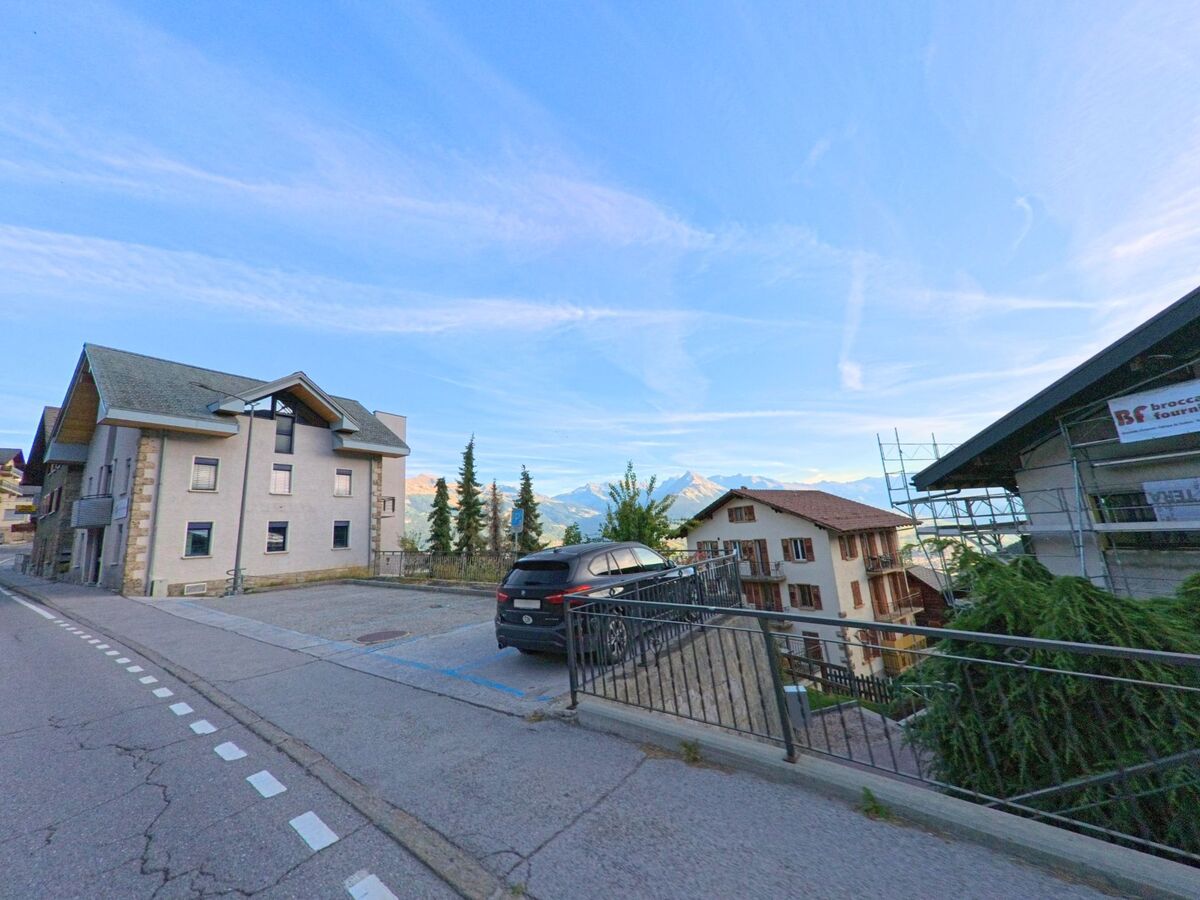 Ein modernes Gebaeude mit Parkplaetzen steht in Basse-Nendaz (1996, Kanton Wallis) neben traditionellen Haeusern mit Blick auf die Berge.