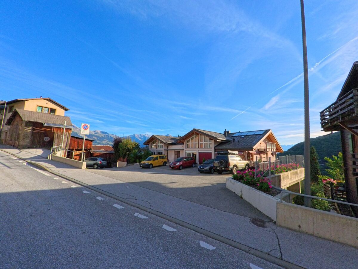Mehrere Wohnhaeuser mit Parkplaetzen liegen in Basse-Nendaz (1996, Kanton Wallis) an einer Strasse mit Blick auf die Berge.