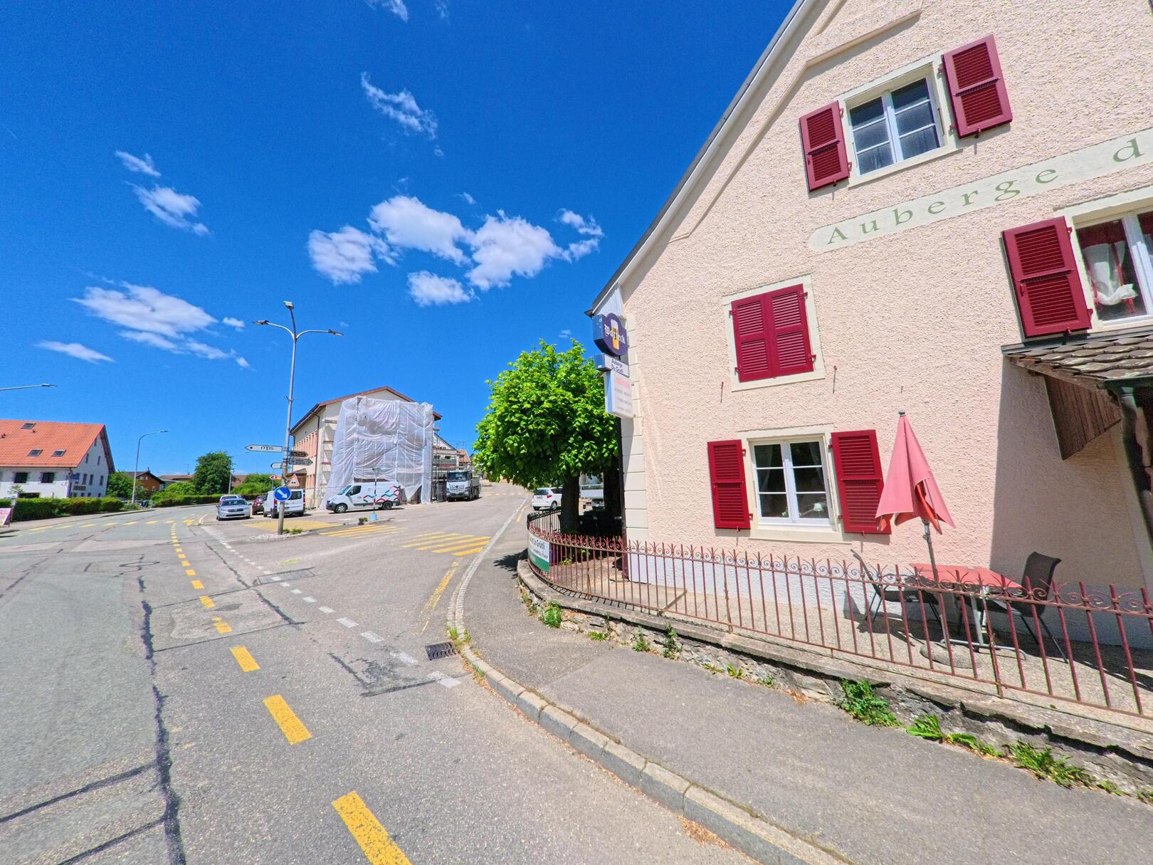 An einer leicht ansteigenden Dorfstrasse in Basse-Vendline (2935, Kanton Jura) steht ein helles Gebäude mit roten Fensterläden unter blauem Himmel.