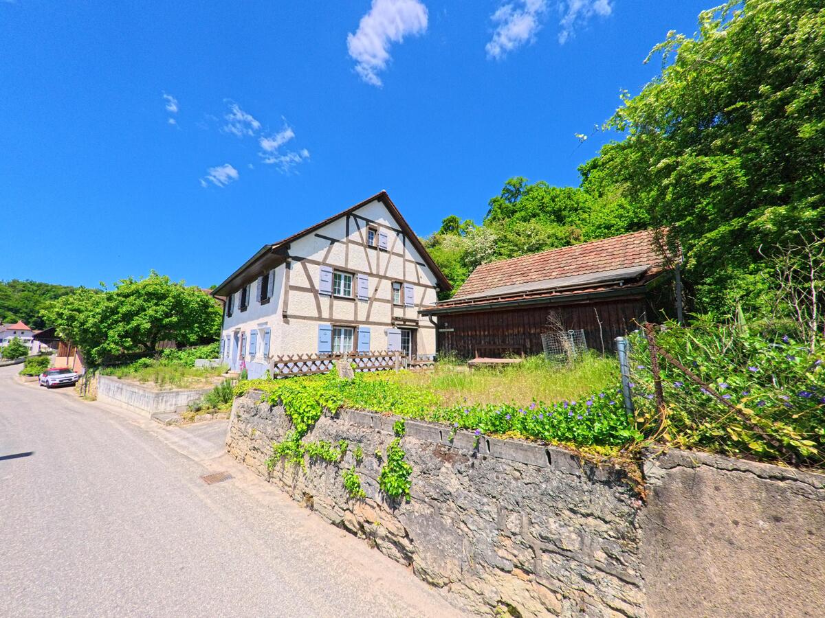 Hinter einer bewachsenen Natursteinmauer steht in Basse-Vendline (2935, Kanton Jura) ein traditionelles Fachwerkhaus mit hellblauen Fensterläden neben einer Holzscheune.