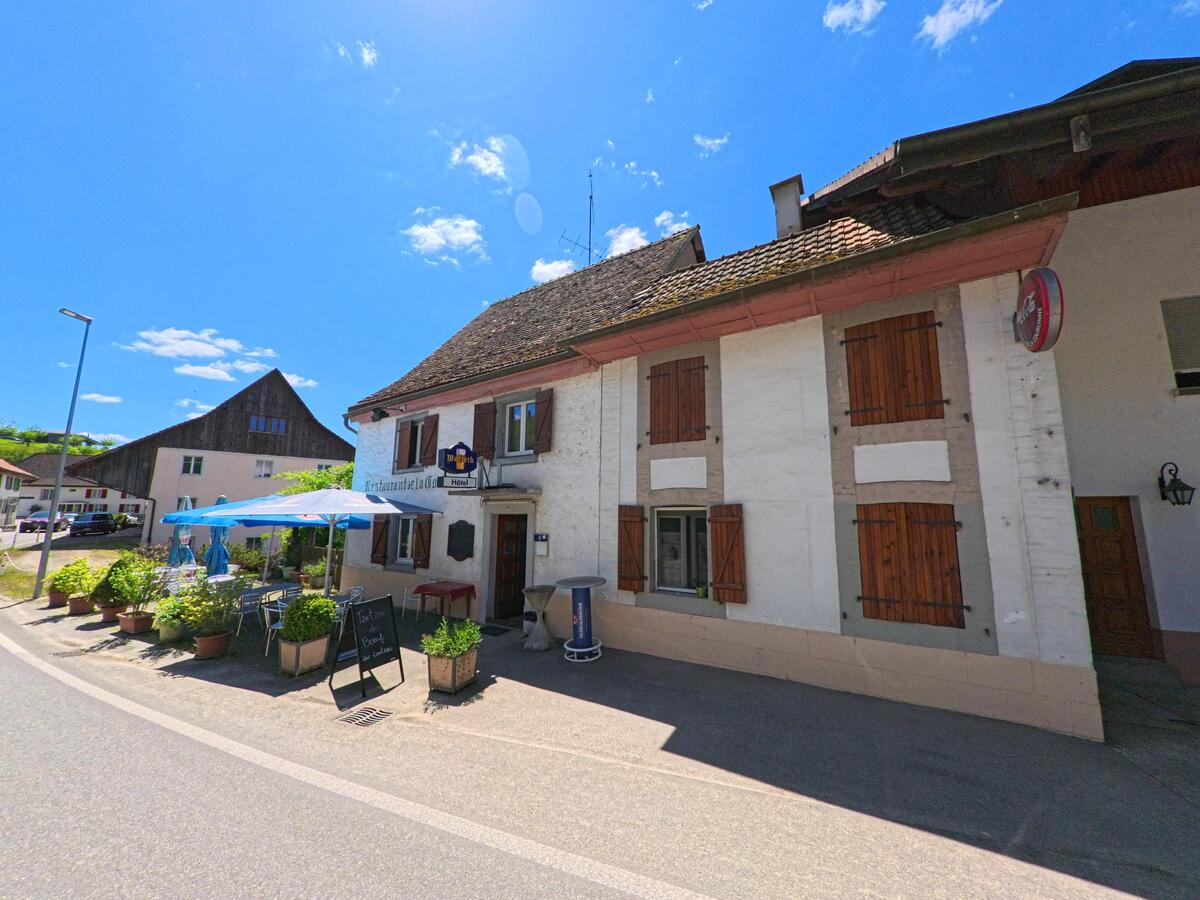 Vor einem traditionellen Gasthaus in Basse-Vendline (2944, Kanton Jura) stehen Tische, Sonnenschirme und bepflanzte Kübel entlang der Strasse.