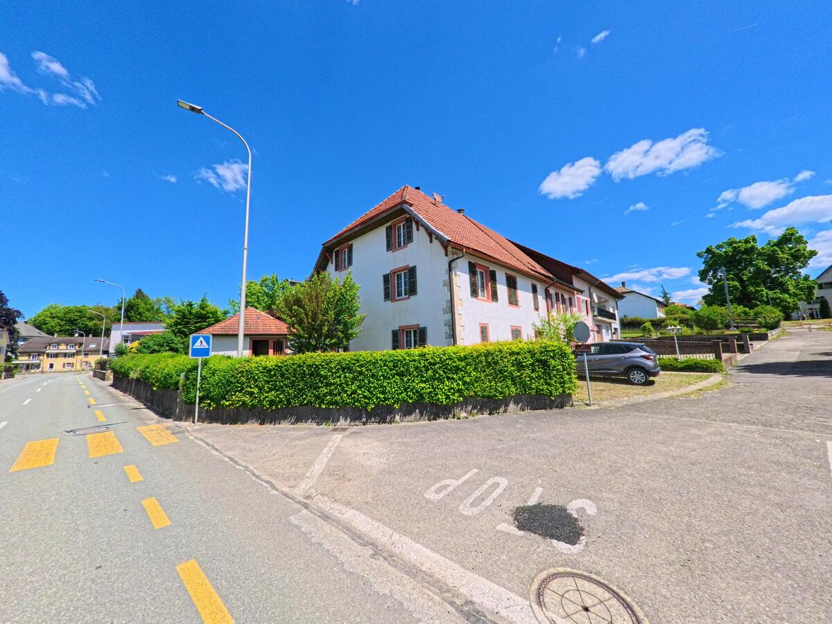 An einer Strassenkreuzung in Basse-Vendline (2944, Kanton Jura) steht hinter einer geschnittenen Hecke ein grosses Gebäude mit rotem Ziegeldach.