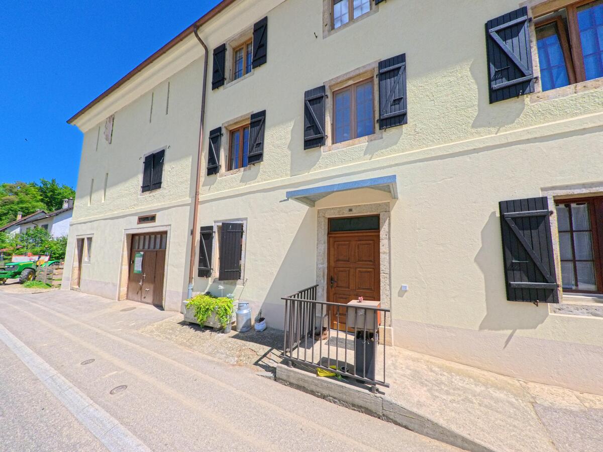 An einer schmalen Dorfstrasse in Basse-Vendline (2944, Kanton Jura) steht ein helles Gebäude mit dunklen Fensterläden und einem hölzernen Eingang.