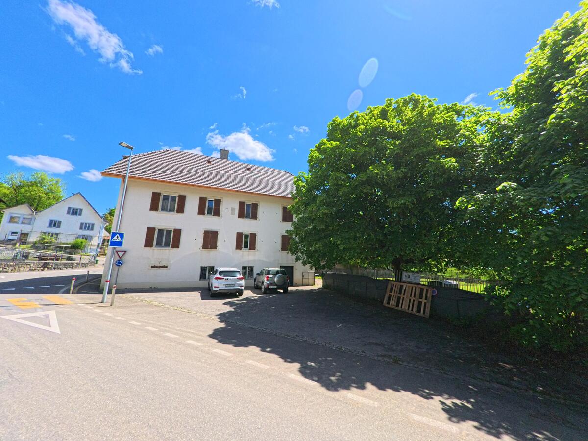 Neben einem Fussgängerstreifen in Basse-Vendline (2944, Kanton Jura) steht ein helles Wohnhaus mit braunen Fensterläden im Schatten grosser Bäume.